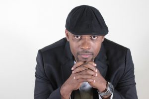 Africian American man, Lasana Omar Hotep, sitting with his hands clutched under his chin looking at the camera wearing a black hat and black suit blazer