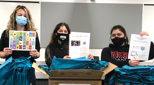 Family Kits Three students wearing non-surgical masks, holding up informational flyers and standing behind cardboard boxes with supplies.