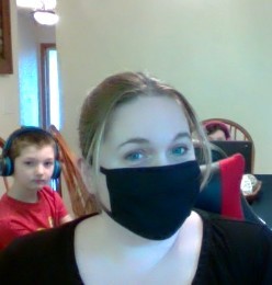 mask selfie - Tracie Tracie Wicks wearing a non-surgical mask with her children sitting at a table behind her. Her children are wearing headphones and have laptops.