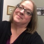 A woman smiles at the camera, she has wire frame glasses and a soft smile. Her head is slightly tilted in the photo.
