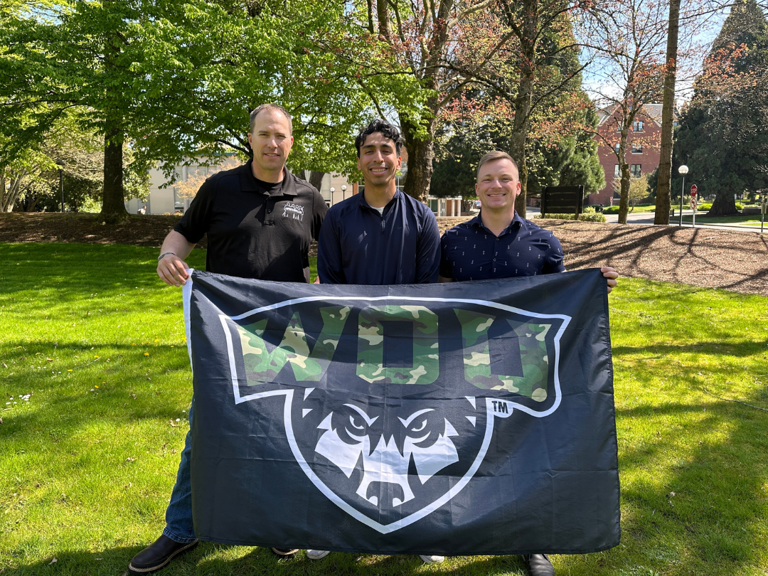 Three men stand on a grassy lawn, holding up a black flag with a "WOU" logo in green camouflage print and a white wolf head. The man on the left wears a black collared shirt and jeans. The man in the middle wears a dark blue long-sleeved shirt and khakis. The man on the right wears a dark blue short-sleeved collared shirt with a subtle pattern and dark pants. Trees and buildings are visible in the background under a clear sky.