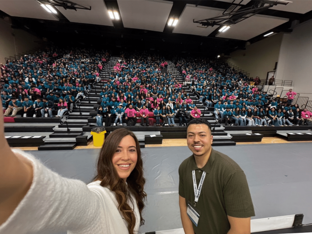 Two smiling individuals pose for a selfie at the 2025 César E. Chávez Leadership Conference. Behind them, a packed auditorium with attendees wearing teal and pink shirts fills the elevated bleacher seating, indicating a large and engaged audience at the event.