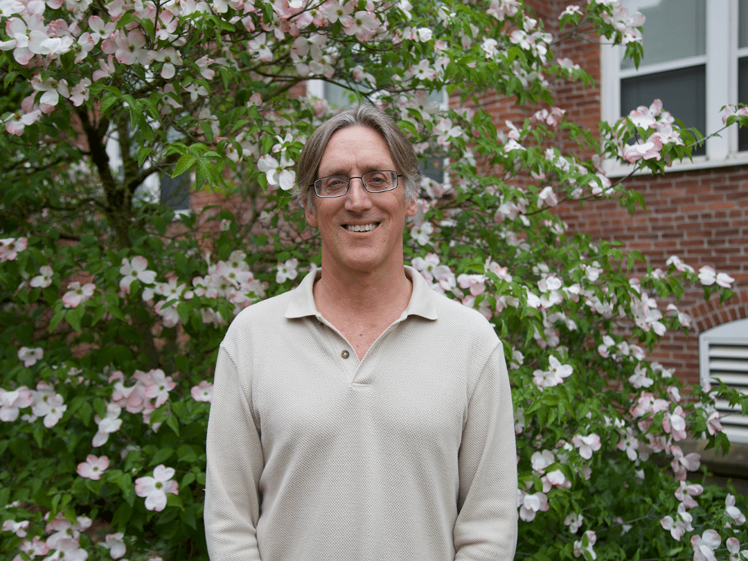 A smiling man with graying hair and glasses stands in front of a blooming dogwood tree and a brick building. He is wearing a light-colored, collared, button-up shirt. The tree behind him is full of white and pale pink flowers and green leaves, creating a soft and colorful backdrop. The red brick of the building is visible in the background, partially obscured by the blossoms. The man is looking directly at the camera with a pleasant expression.