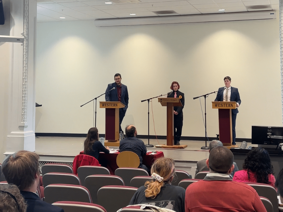 Image of 3 students on a stage with mics presenting to a group of people