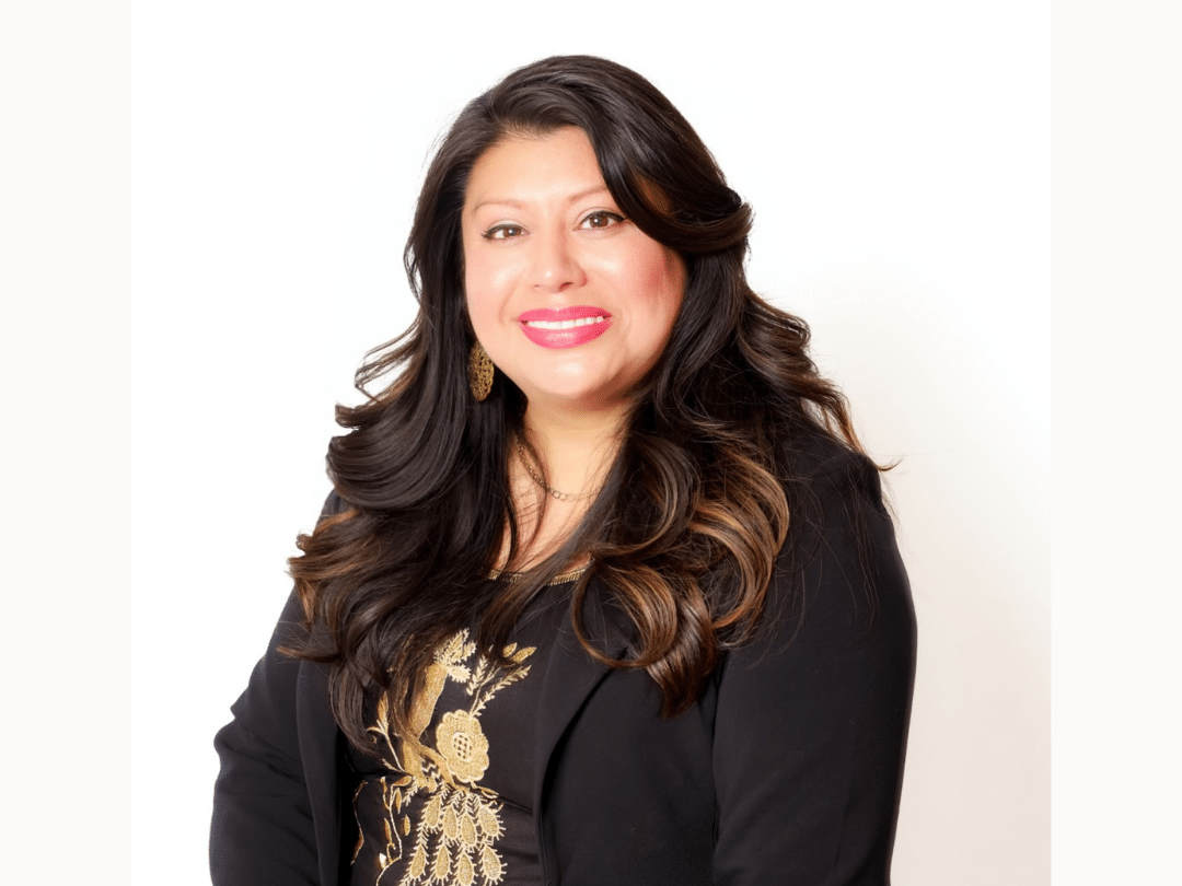 A smiling woman with long, dark, wavy hair, wearing a black blazer over a dark top with gold embellishments, and gold hoop earrings, poses against a plain white background.