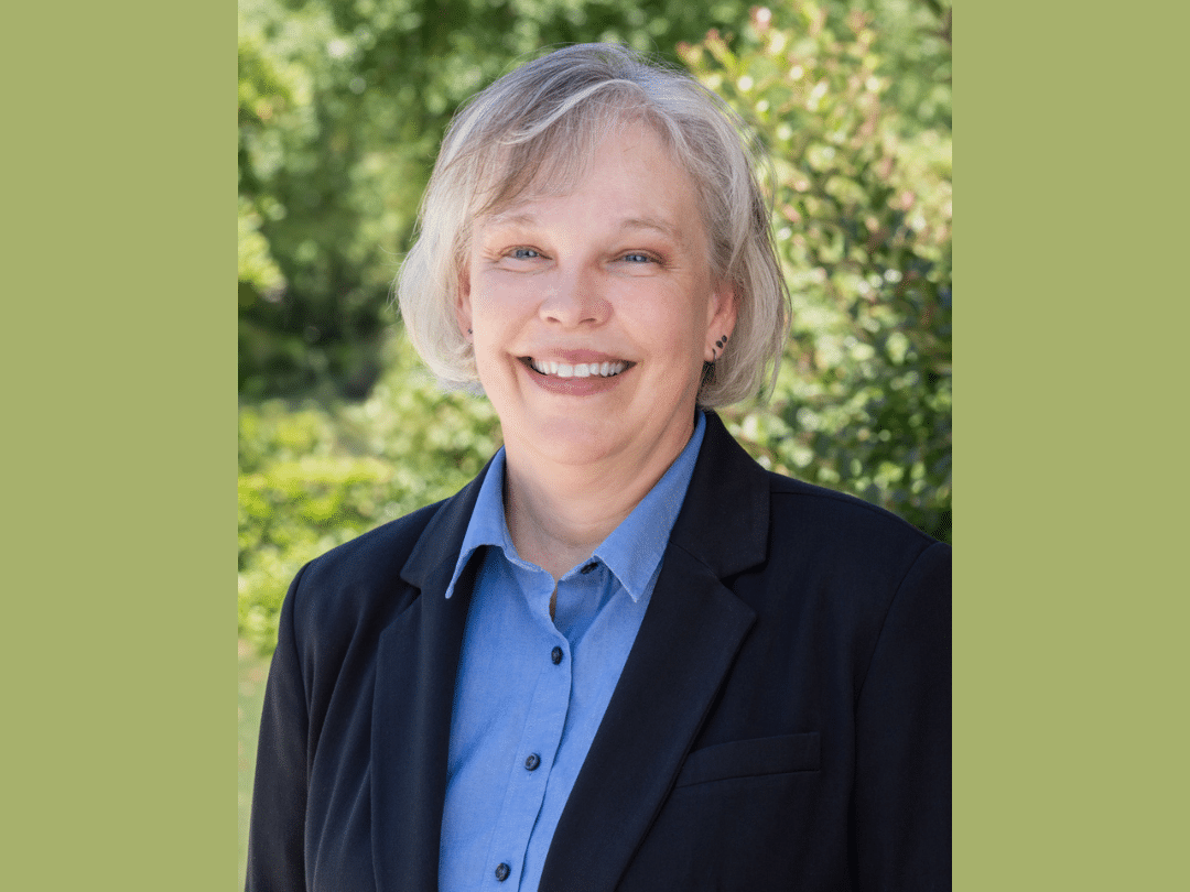 A headshot of a smiling woman with short, gray hair and fair skin. She is wearing a black blazer over a light blue collared shirt. She has small earrings and appears to be outdoors with green foliage in the blurred background.