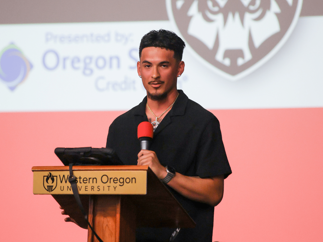 Athletic Auction PR A man with short dark hair and a beard, wearing a black collared shirt and a necklace, stands at a wooden podium with 'Western Oregon University' inscribed on it. He holds a microphone in his right hand and looks forward. Behind him is a screen displaying 'Presented by: Oregon S Credit' and a stylized animal head logo.