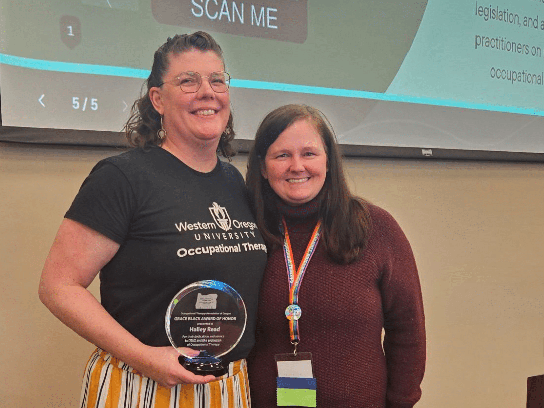 A smiling woman in a black t-shirt and striped pants holds a clear award plaque that reads "Grace Black Award of Honor, Halley Read." She stands next to another smiling woman in a maroon sweater with an ID badge around her neck. In the background, a screen displays text and a "SCAN ME" QR code.